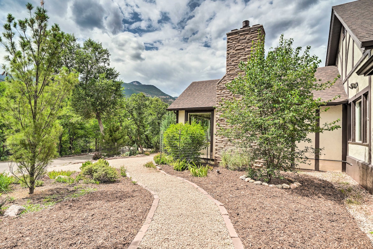 A landscaped outdoor area features a winding gravel pathway leading to the entrance of a home. Surrounding the pathway are various shrubs and trees, with a prominent stone chimney visible on the facade of the building.