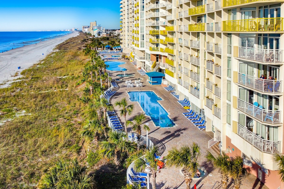 The image captures the expansive outdoor pool area, lined with lounge chairs, set against the backdrop of the beach and ocean. Lush palm trees provide a natural touch, and the building's balconies are visible, each adorned with yellow accents. Clear skies enhance the inviting atmosphere.