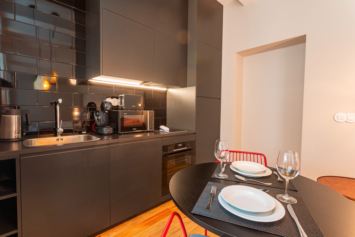 A modern kitchen area is presented, featuring sleek black cabinetry and stainless steel appliances. A round table is set with dishes and glassware, providing a dining space. Soft lighting creates a warm ambiance, enhancing the contemporary design elements.