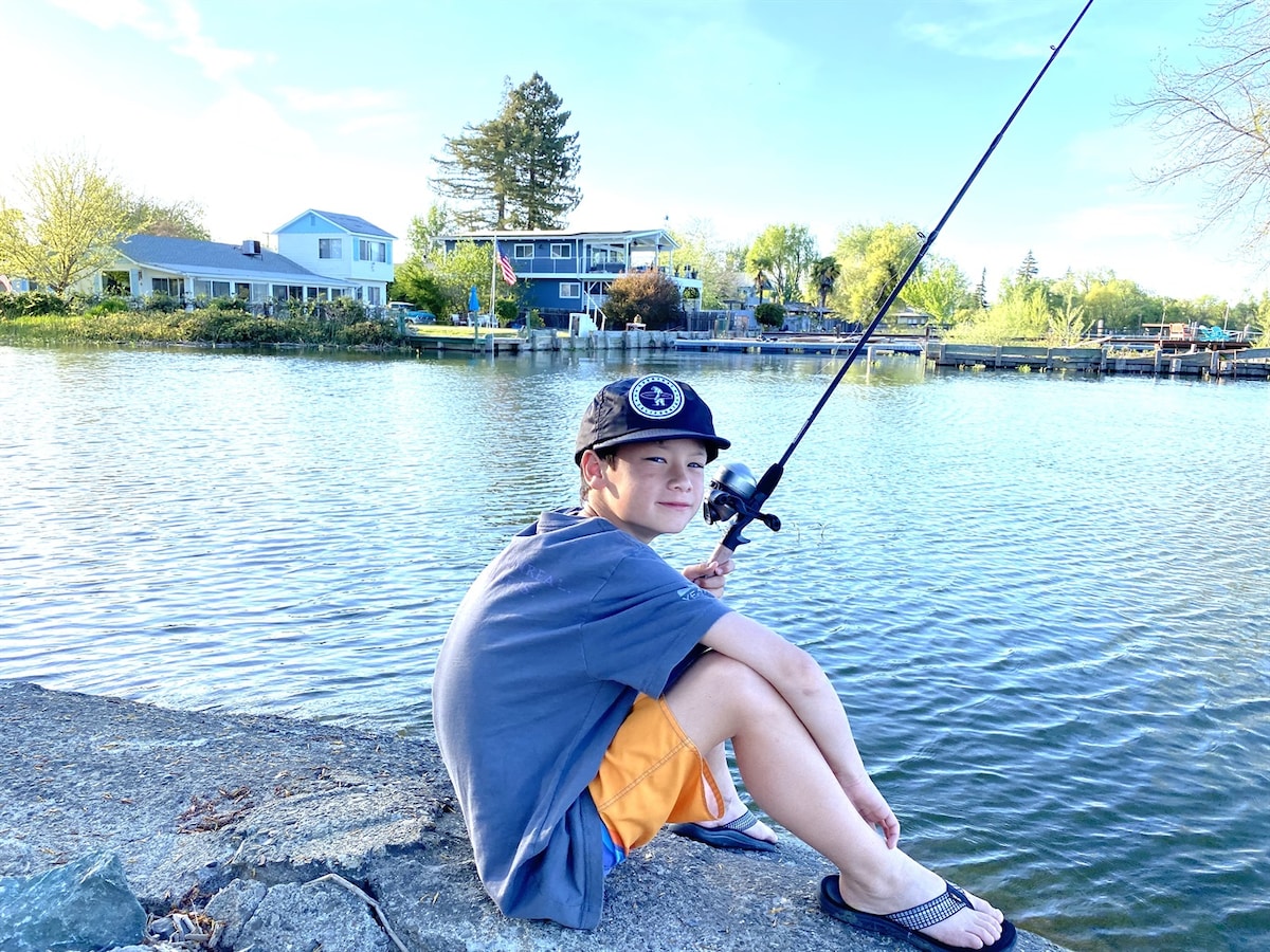 A child sits on a rock by the water, focused on fishing with a rod. The calm lake is visible in front, framed by greenery and distant homes. Soft sunlight illuminates the scene, creating a peaceful outdoor setting.