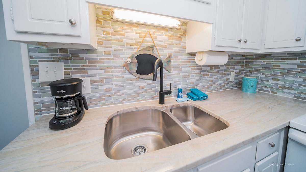A modern kitchen features a double-basin sink set into a light-colored countertop. A coffee maker is positioned nearby, and a decorative fish hangs on the wall above the sink. Soft blue and gray tiles create a calming backsplash, complemented by neatly arranged kitchen essentials.