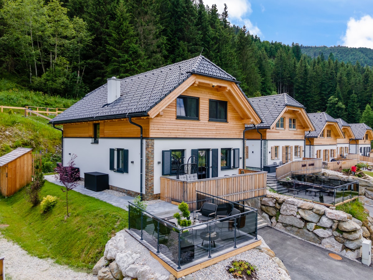 A modern chalet is situated amidst lush greenery, featuring a wooden façade with large windows and a sloped roof. A glass railing surrounds a patio area furnished with a small table and chairs, offering views of the natural surroundings.