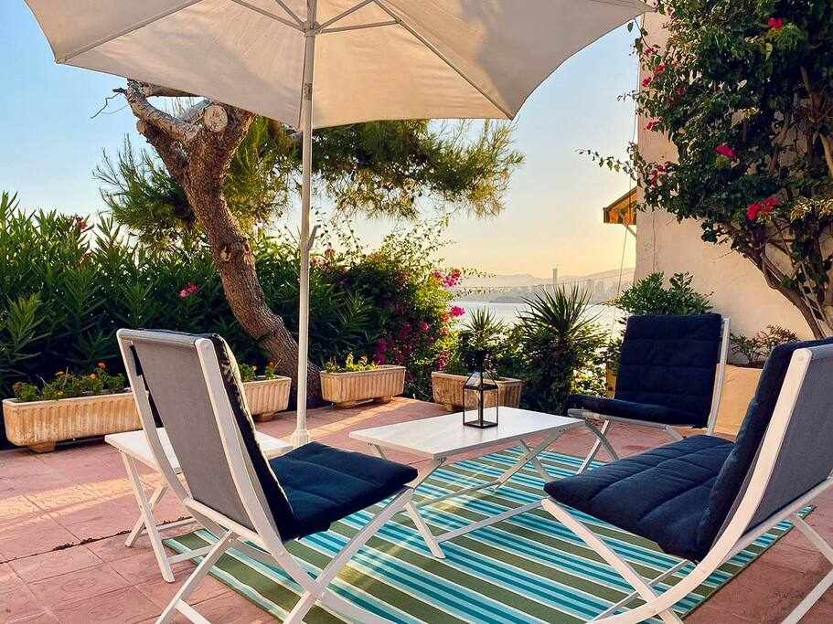 An outdoor patio area features two lounge chairs and a small table, positioned under a large umbrella. Lush plants provide privacy, with vibrant greenery framing the view. A striped rug adds color beneath the seating arrangement, creating a relaxing space for enjoyment.