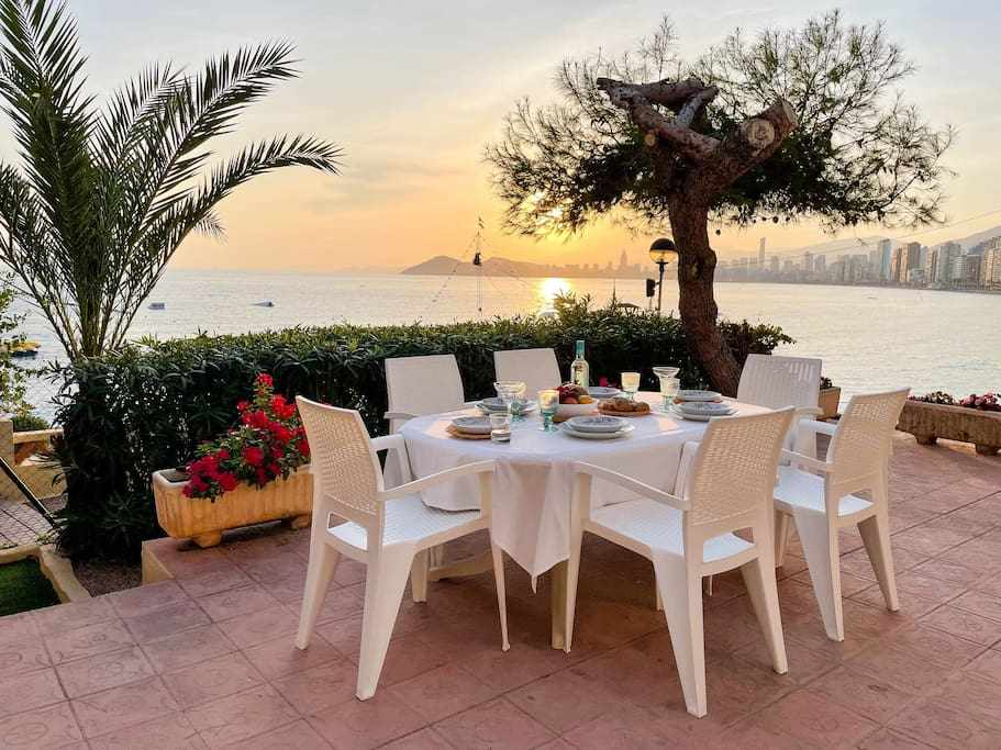 An outdoor dining area is set for six, featuring a white table draped with a tablecloth. The backdrop reveals a serene sunset over the sea, with distant city skyline and boats on the water. Lush greenery and colorful flowers surround the space.