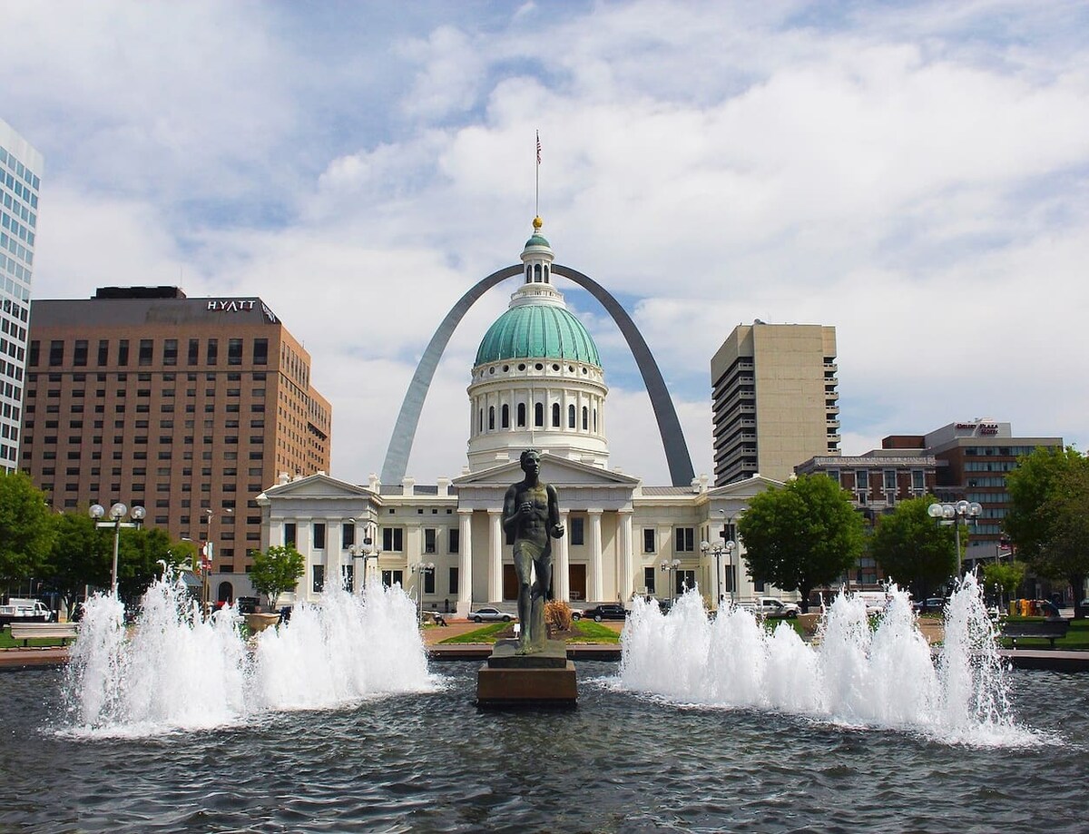 A lively fountain is featured in the foreground, with jets of water cascading down. The iconic Gateway Arch stands prominently in the background, along with the historic Old Courthouse, which is topped with a green dome. Surrounding buildings showcase urban architecture.