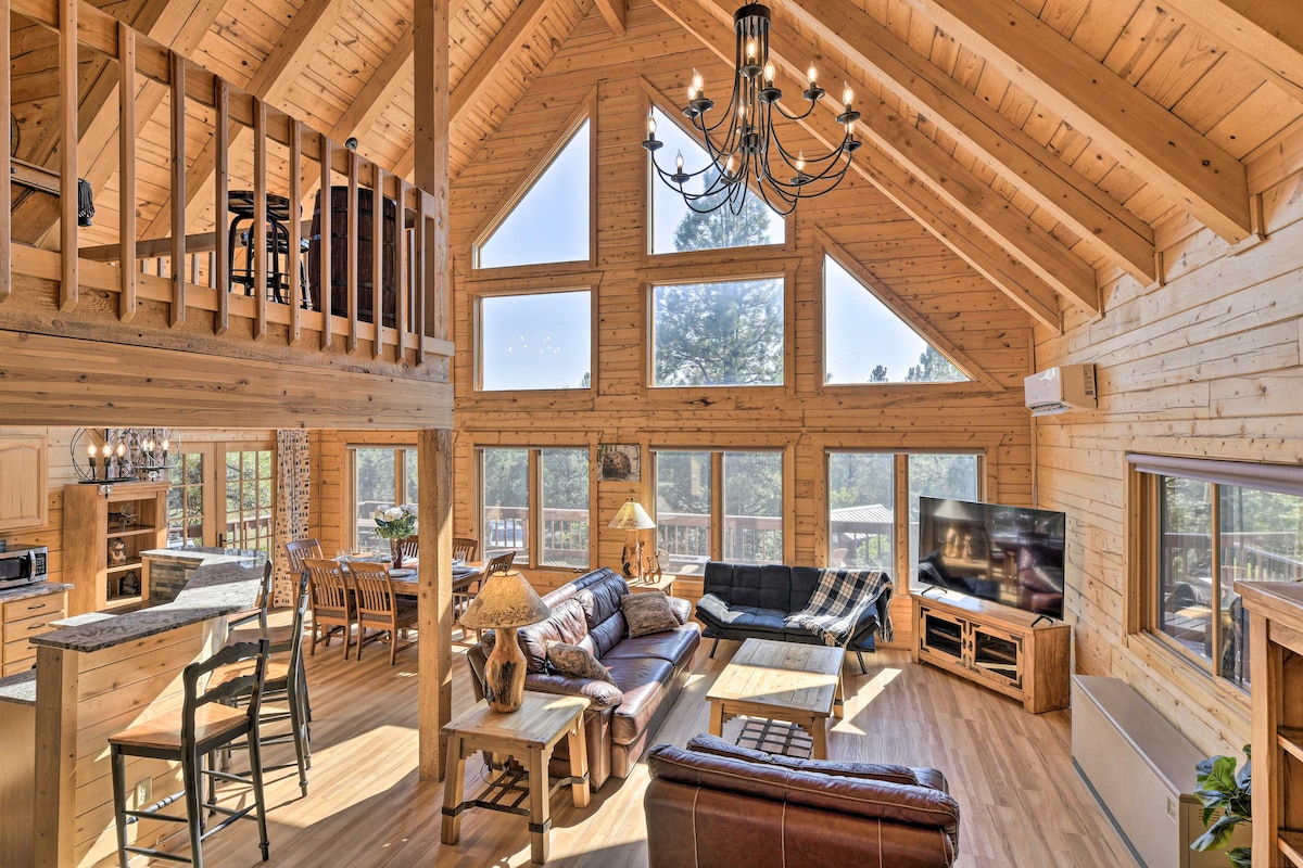 The spacious living area features a high, A-frame ceiling with exposed wooden beams. Large windows provide ample natural light and views of the surrounding forest. Comfortable seating surrounds a coffee table, with a dining area and access to the upstairs loft visible in the background.