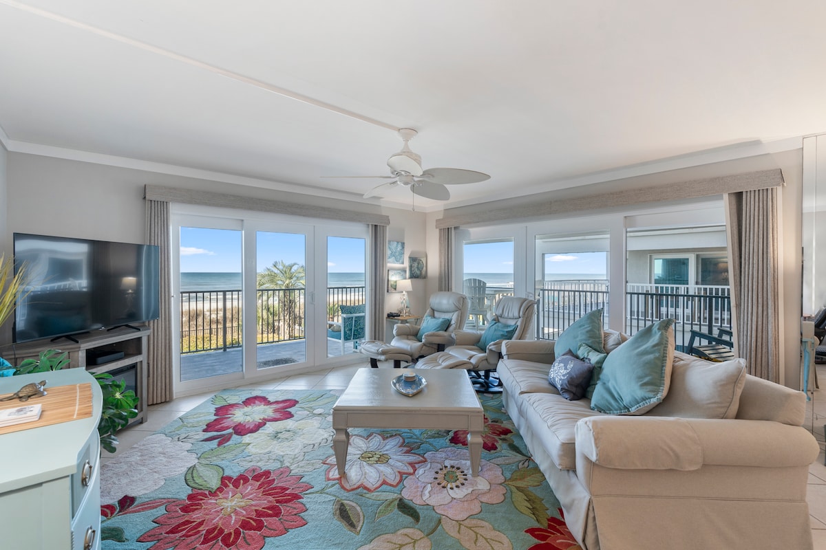 An inviting living area features large glass doors that open to ocean views. Comfortable seating is arranged around a central coffee table, accented by a patterned area rug. Light pours in from the windows, enhancing the spaciousness of the room.