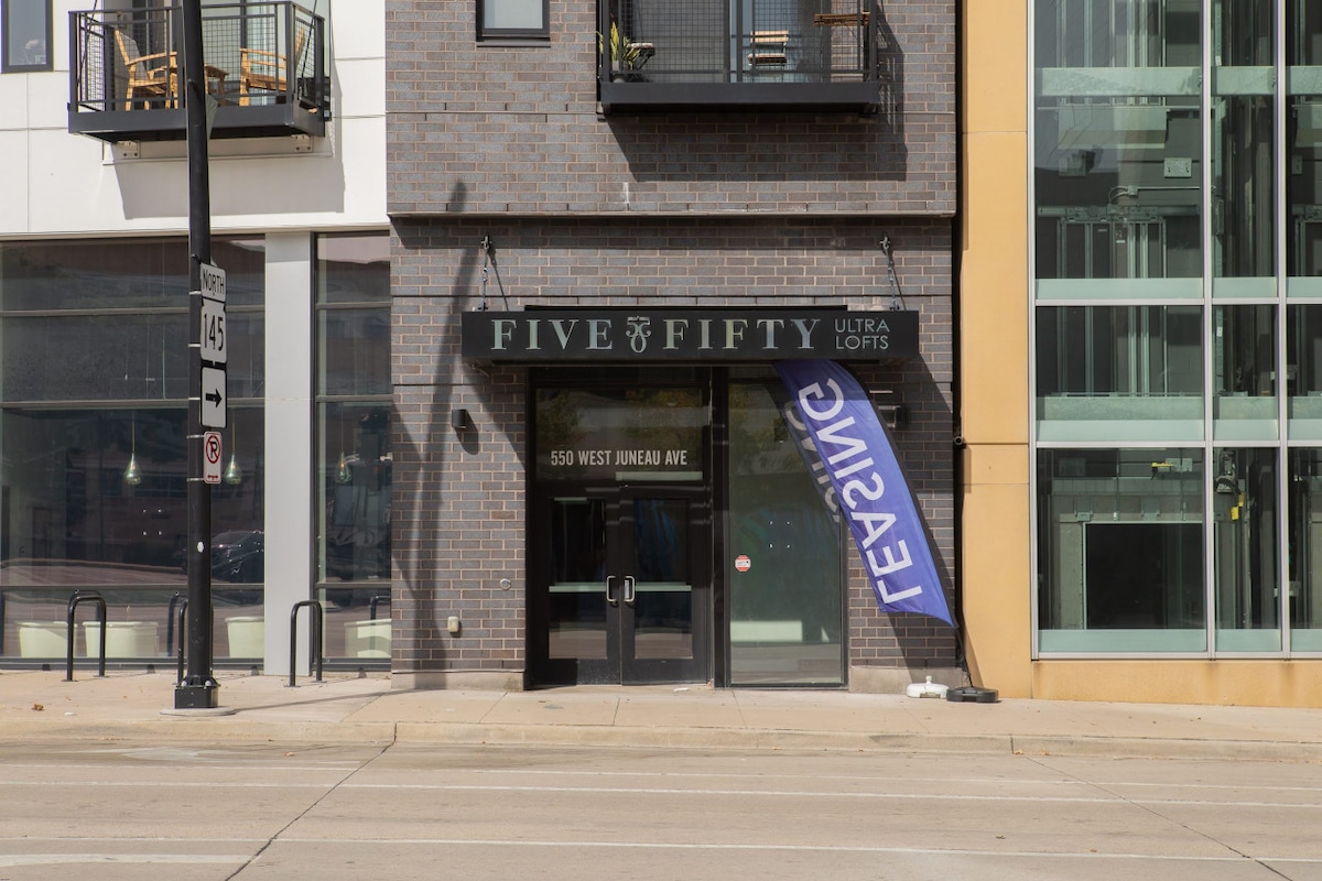 The image shows the entrance of the Five Fifty Ultra Lofts building, featuring a glass door with the address displayed above. A blue leasing banner is positioned to the right, and large windows of the building are visible on the right side.