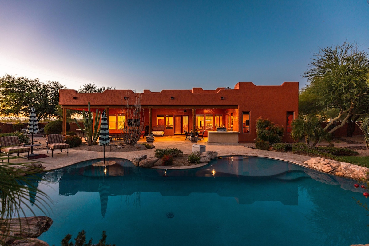 The mansion's exterior is illuminated during twilight, showcasing warm tones against a dusky sky. A sparkling resort-style pool reflects the evening light, surrounded by landscaped gardens and seating areas, inviting relaxation and outdoor gatherings.