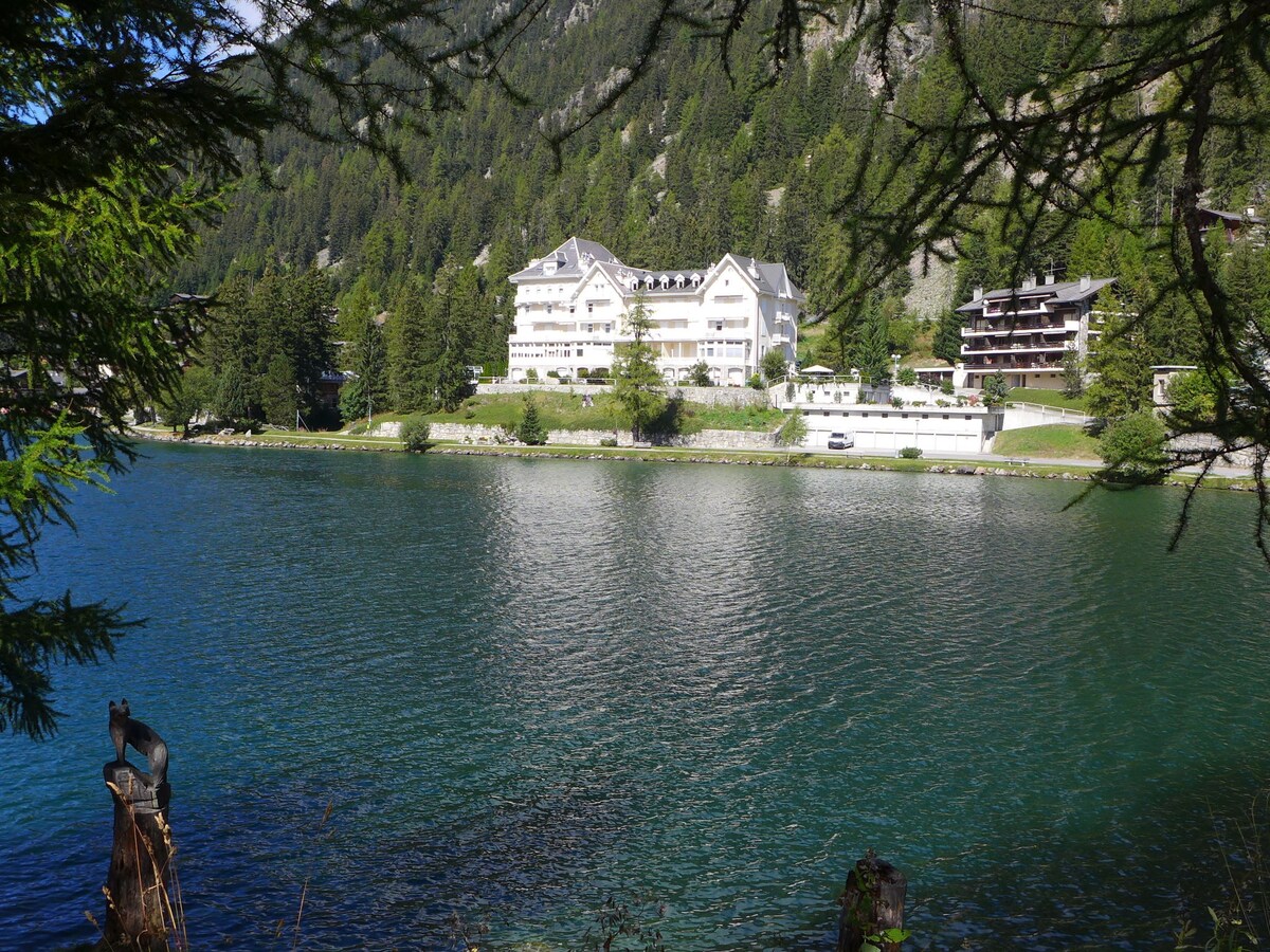 A view of the apartment house 'Alpes et Lac' set against a backdrop of mountains and trees. The lake is visible in the foreground, reflecting the greenery and buildings. The calm water creates a serene atmosphere amid nature.