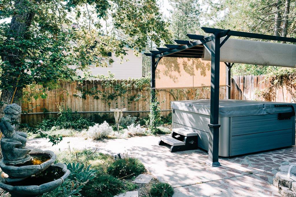 A private outdoor area features a hot tub under a shaded pergola, surrounded by lush greenery and a stone pathway. A decorative fountain is positioned nearby, adding a tranquil element to the space, while natural light filters through the trees.