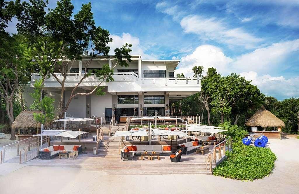 A spacious outdoor area is featured, showcasing comfortable seating arrangements beneath large umbrellas. The structure behind includes large windows and multiple levels, surrounded by lush greenery. Beach equipment, including blue kayaks, is positioned nearby, highlighting the resort's proximity to the waterfront.