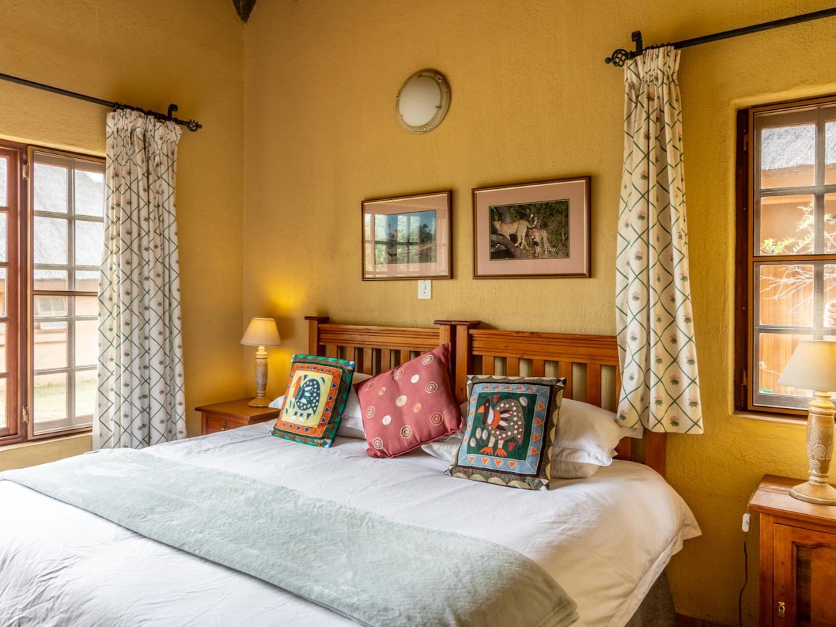 A cozy bedroom features a spacious bed adorned with colorful decorative pillows. Soft linens and a light blanket enhance the inviting feel. Two bedside lamps provide warm illumination, while framed art and light curtains complement the natural light streaming through the windows.