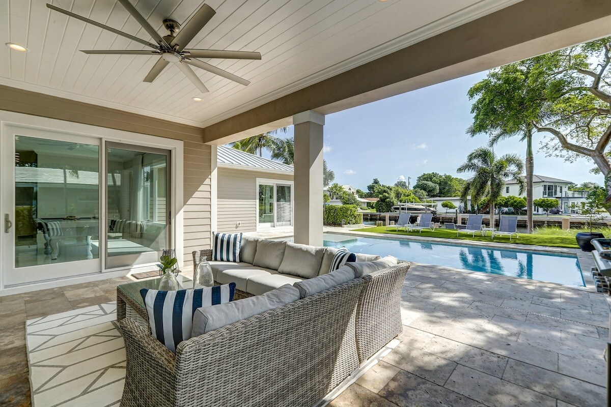 A covered patio area features a comfortable sectional sofa adorned with blue and white striped cushions. The space is complemented by a ceiling fan and overlooks a serene pool surrounded by lush greenery and homes beyond.
