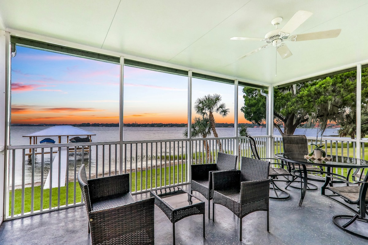 A screened-in porch provides an unobstructed view of Dinner Lake, framed by lush greenery. Comfortable seating is arranged around a round table, with the warm hues of sunset visible through expansive windows. A ceiling fan offers a gentle breeze, enhancing the relaxing atmosphere.