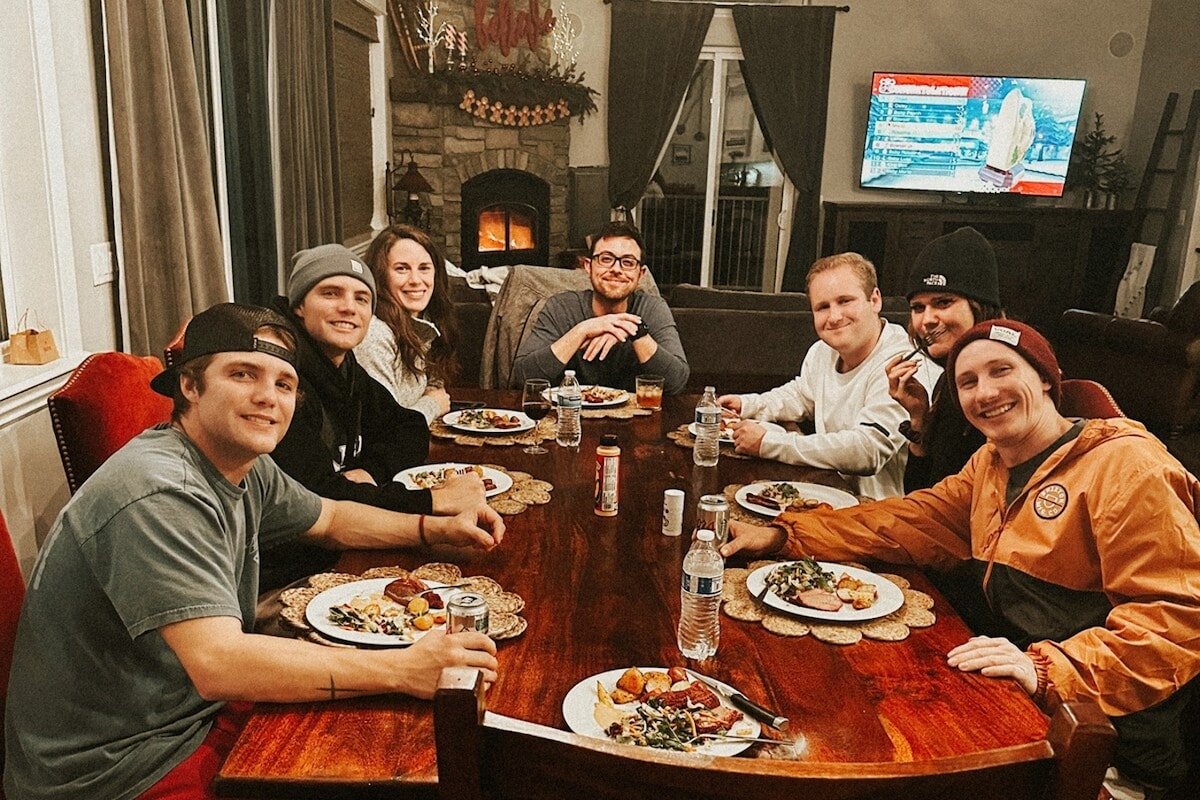 A spacious dining table is surrounded by guests enjoying a meal. Various plates filled with food are present, with water bottles and soft drinks also on the table. A cozy fireplace can be seen in the background, alongside a flat-screen TV displaying a news program.
