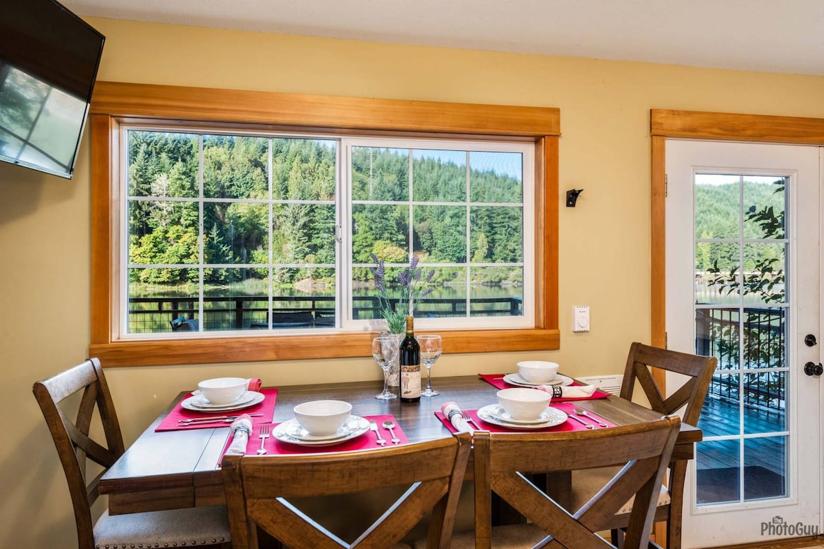 A dining table set for four is positioned near a large window, offering views of lush greenery. Natural light fills the space, and table settings include white plates and glassware. A door leads to the outdoor area, enhancing the inviting atmosphere.