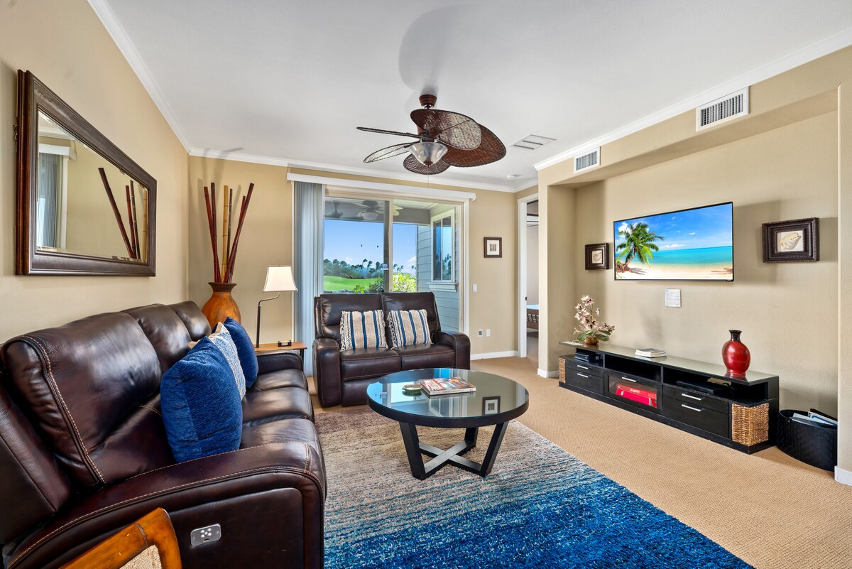 The living area features a combination of comfortable brown leather sofas and a round glass coffee table, set against a backdrop of soft beige walls. A flat-screen TV is mounted on the wall, with a view of the greenery outside through sliding glass doors, providing natural light.