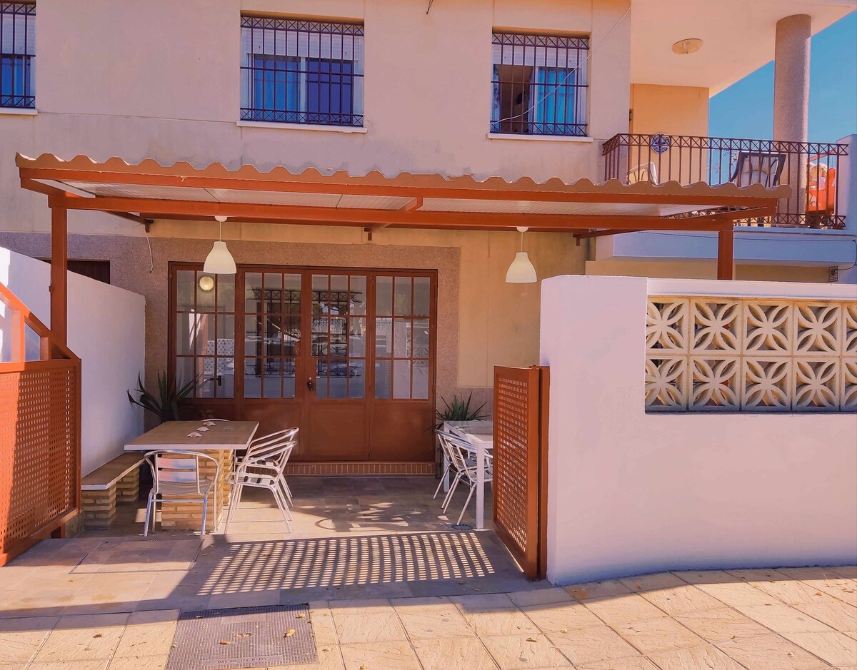 An outdoor patio area is showcased, featuring a wooden structure with a roof overhead. Tables and chairs are arranged to encourage relaxation. The walls are decorated with geometric patterns, and large windows provide light to the adjacent indoor space.