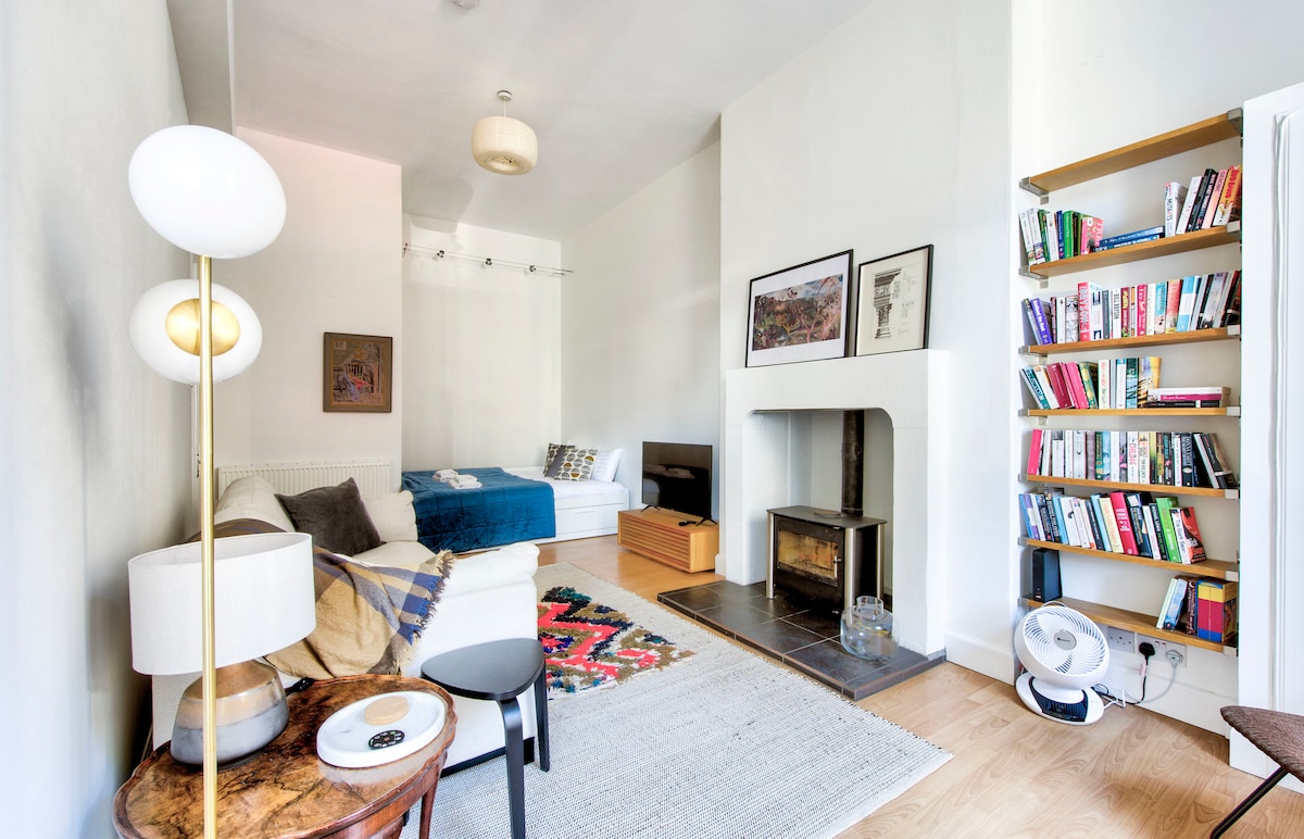 A modern living room features a comfortable sofa adorned with patterned cushions and a stylish rug. A bookshelf filled with books is visible, alongside a fireplace. A cozy sleeping area is partially seen in the background, with a double bed made with fresh linen.
