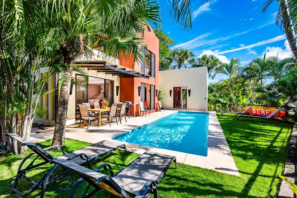 A modern home is framed by lush tropical gardens, featuring a clear private pool surrounded by sun loungers. The patio area showcases a dining set, ideal for outdoor meals. The structure's multi-story design allows for ample natural light to illuminate the open spaces.