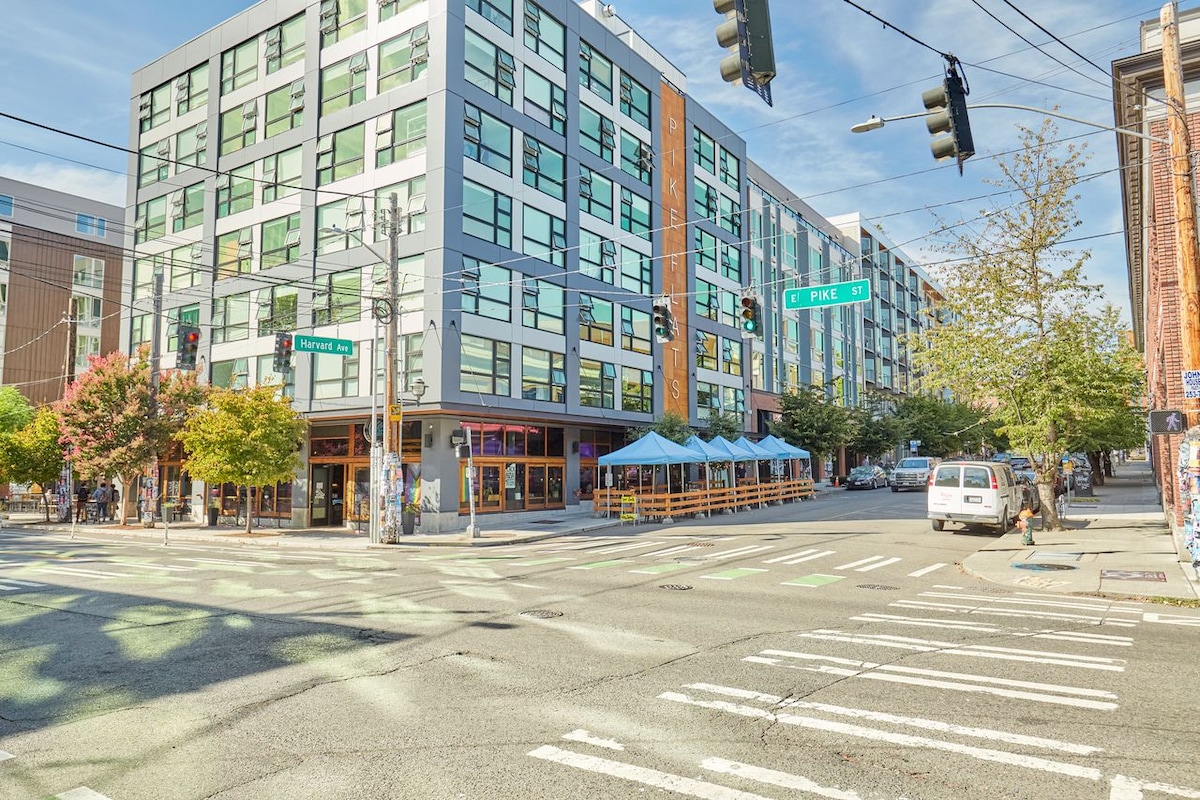 A modern multi-story building is set at the intersection of two streets, with large windows reflecting the ample daylight. Trees are seen lining the sidewalks, and outdoor seating can be noted in front of a café. Traffic signals and parked cars are visible.