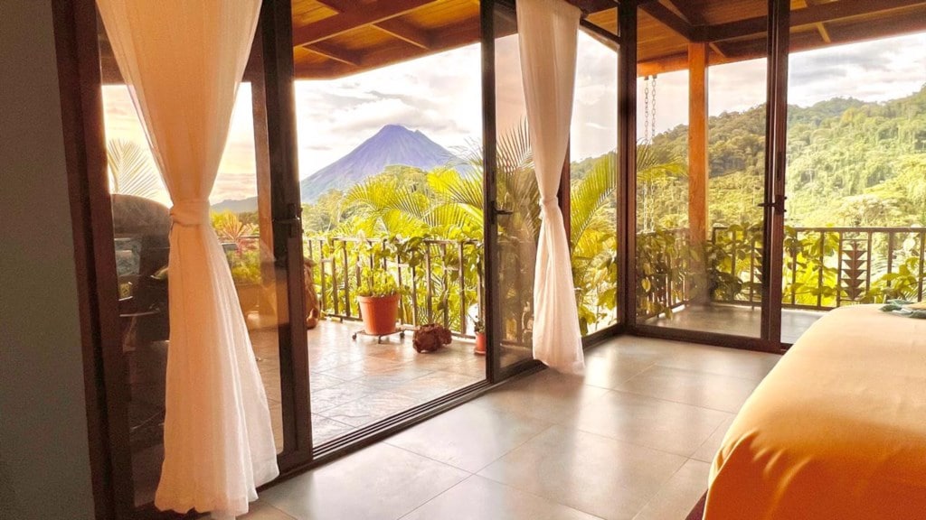 The spacious room features floor-to-ceiling glass walls, providing an unobstructed view of Arenal Volcano and lush, green surroundings. Natural light fills the space, and flowing white curtains frame the entrance to a private terrace with tropical plants and a cozy atmosphere.