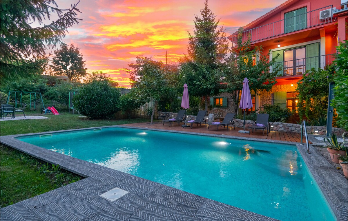 A swimming pool is framed by a wooden deck and lounge chairs, set against a vibrant sunset sky. Trees surround the area, adding greenery, while the colorful façade of the house is visible in the background. Play equipment can be seen on the lawn.
