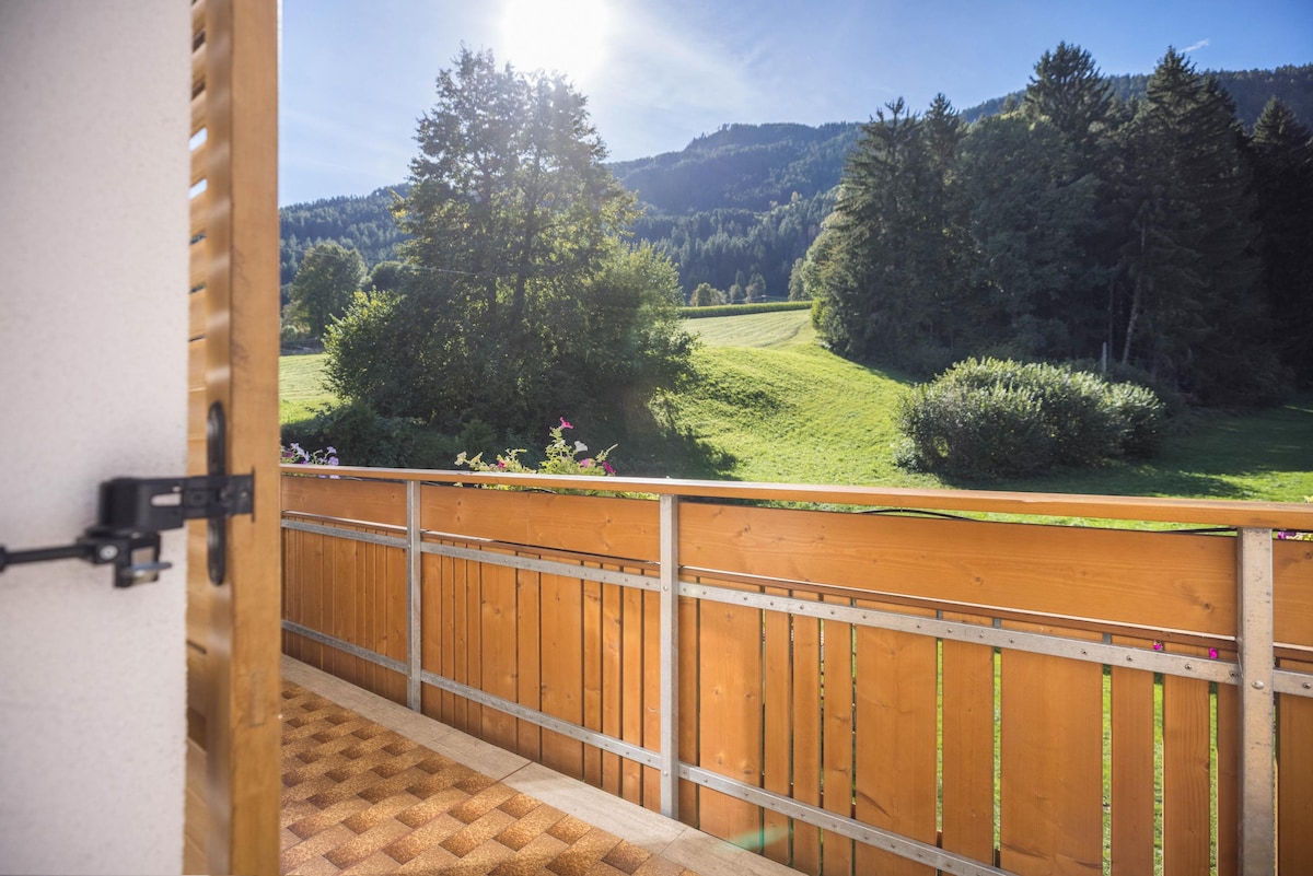 A wooden balcony offers a view of lush green hills and trees under a clear blue sky. Sunlight floods the scene, creating a bright outdoor space. The balcony's railing features vertical wooden slats, with flower boxes visible, adding a touch of color to the setting.