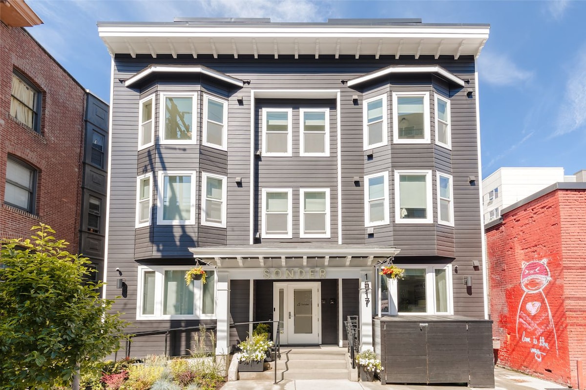 The exterior of a three-story building features a contemporary design with multiple large windows. A landscaped area with flowers is visible in front, enhancing the building's curb appeal. The entrance is clearly marked and accessible, showcasing a welcoming atmosphere.