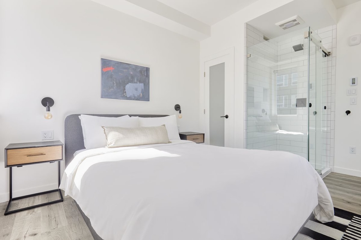 A bright and airy bedroom features a spacious bed with crisp white linens. Two bedside tables are positioned on either side, each equipped with stylish wall-mounted lamps. A glass shower area is visible in the corner, complemented by modern tile work.