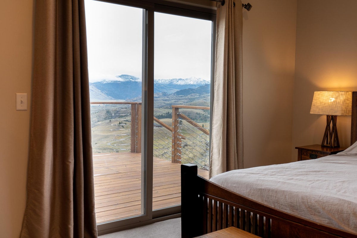 A bedroom features a spacious bed with light bedding, framed by floor-to-ceiling windows that offer stunning views of the surrounding mountains. Sliding glass doors lead to an expansive wooden deck, enhancing the connection to the picturesque landscape outside.