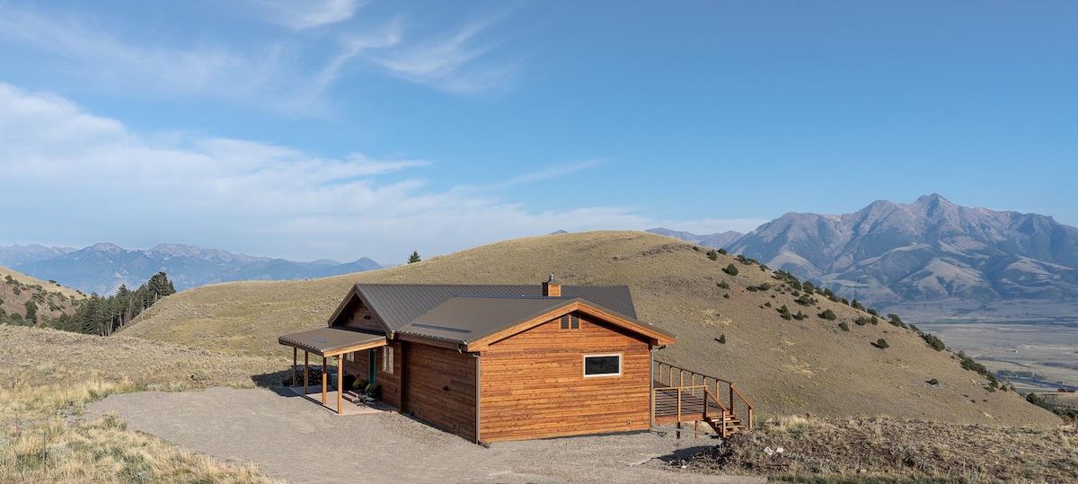 A wooden mountain retreat is captured on a hillside, framed by the Absaroka Mountains in the background. The home features a welcoming porch and large windows, blending harmoniously with the surrounding natural landscape. Clear skies and rolling hills create an expansive atmosphere.