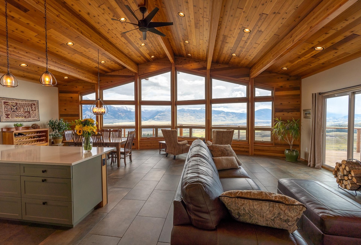 An open-concept living space features vaulted wooden ceilings and large picture windows showcasing scenic mountain views. A sectional sofa and stylish accent pillows provide seating, while a dining area with wood chairs and a table complements the rustic decor. A modern ceiling fan enhances airflow.