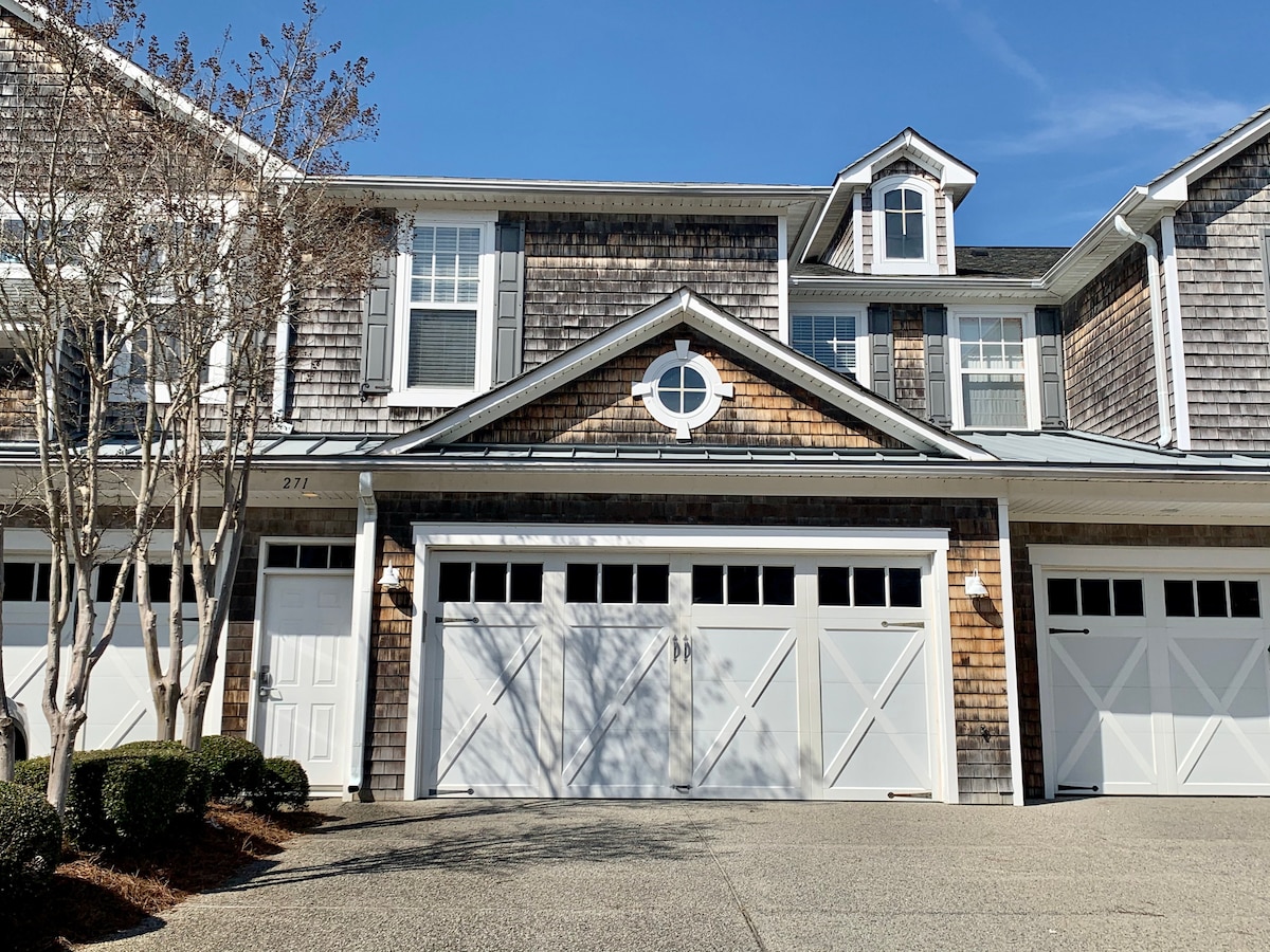 A charming exterior view of a multi-story home features shingle siding and multiple gabled roofs. The garage contains three doors with decorative cross patterns. Sunlight casts shadows on the neatly landscaped front yard, which includes trimmed shrubs and a concrete driveway.