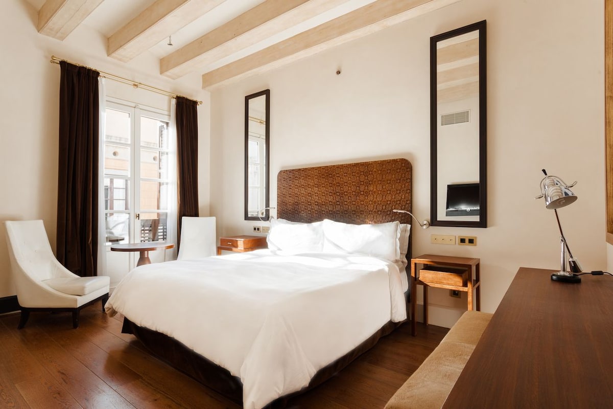 A spacious room features a large bed with a woven headboard and natural light filtering through the window. Two mirrors are mounted on the walls, reflecting the minimalist wooden furnishings. A comfortable chair and desk are positioned beside the bed, enhancing the room's inviting feel.