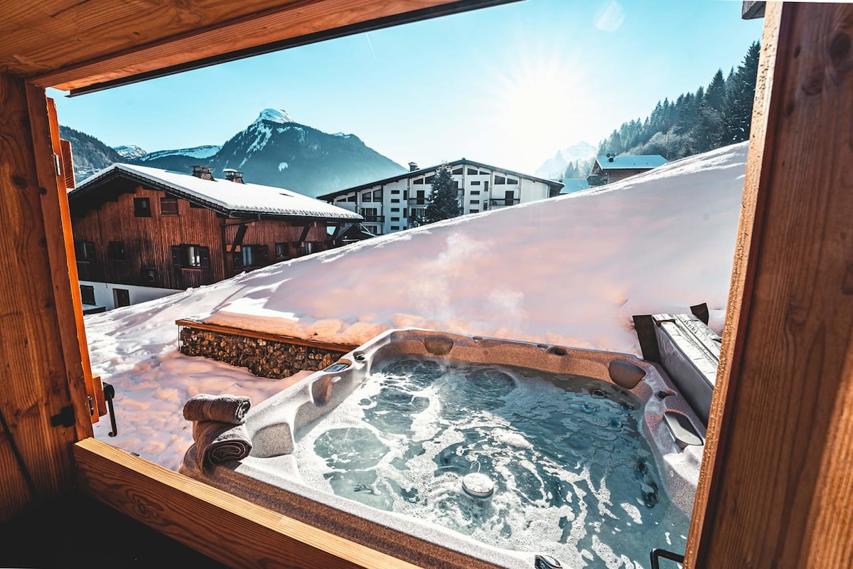 A hot tub is positioned on a private terrace, with snow-covered mountains and clear blue skies visible in the background. The serene landscape features a variety of alpine buildings, creating a tranquil setting that invites relaxation.