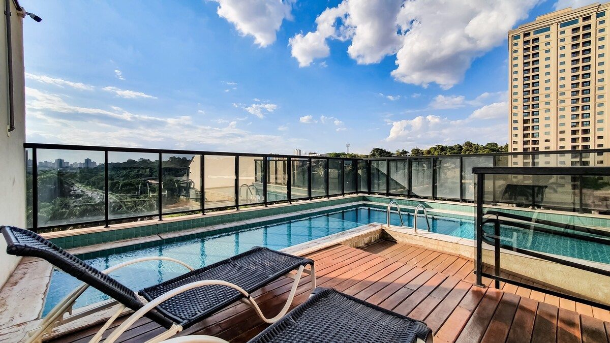 A rooftop pool area is displayed, featuring a clear blue water surface and surrounding glass panels. Lounge chairs are positioned nearby, offering a space to relax and enjoy the expansive sky filled with clouds. The city skyline is visible in the background.