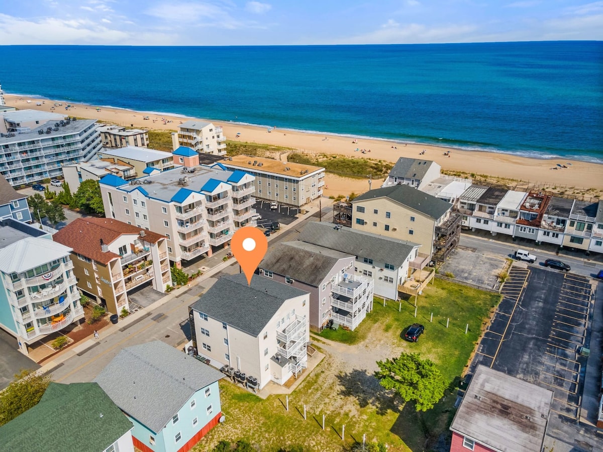 An aerial view captures the Seaport building on a block near the beach, showcasing the sandy shoreline and serene ocean waters. Surrounding buildings and parking areas are visible, highlighting the unit's proximity to local amenities and the beach.