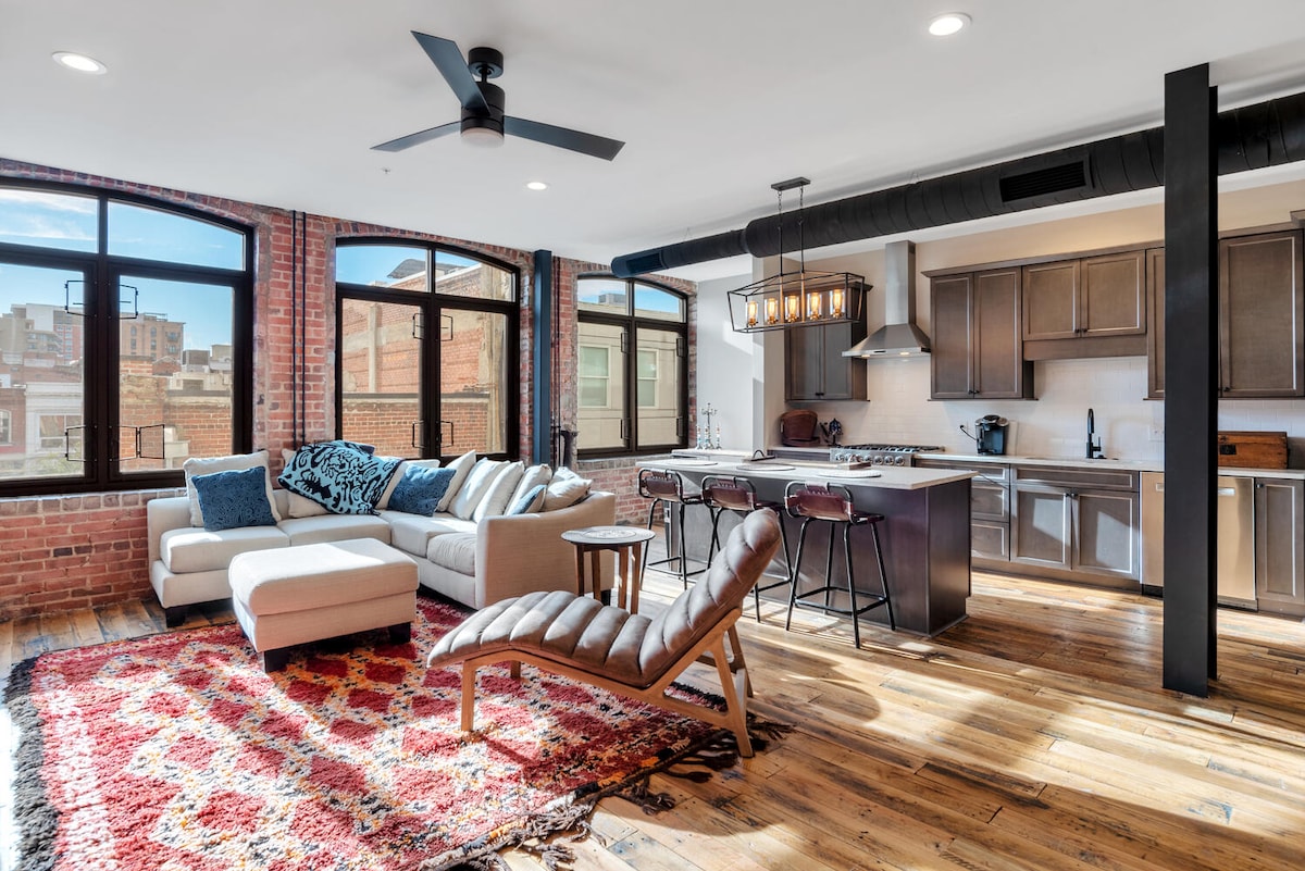 A spacious living area is highlighted by large windows that provide ample natural light, revealing exposed brick walls and wooden flooring. A modern sectional sofa and a lounge chair are arranged on a vibrant area rug, while a kitchen area features a central island with pendant lighting.