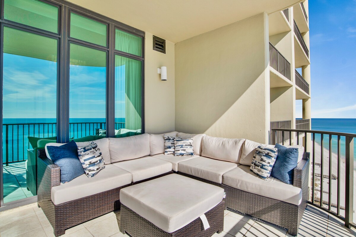 An outdoor living space features a large sectional sofa in neutral tones, complemented by blue and white accent pillows. Behind, floor-to-ceiling windows allow for unobstructed ocean views, while a gently tiled floor extends toward the balcony railing.