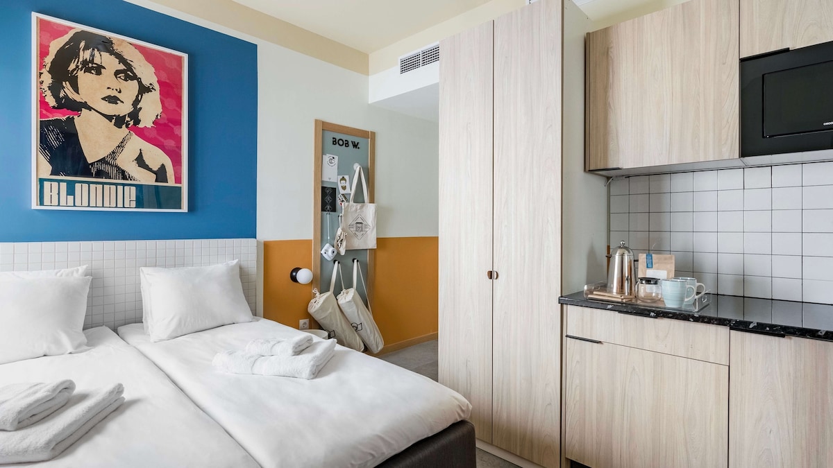 The studio features a comfortable sleeping area with two beds arranged side by side. A kitchenette is visible with essential appliances and dishware. Colorful wall art adds visual interest, while soft lighting enhances the overall ambiance. Storage space is also available beside the kitchenette.