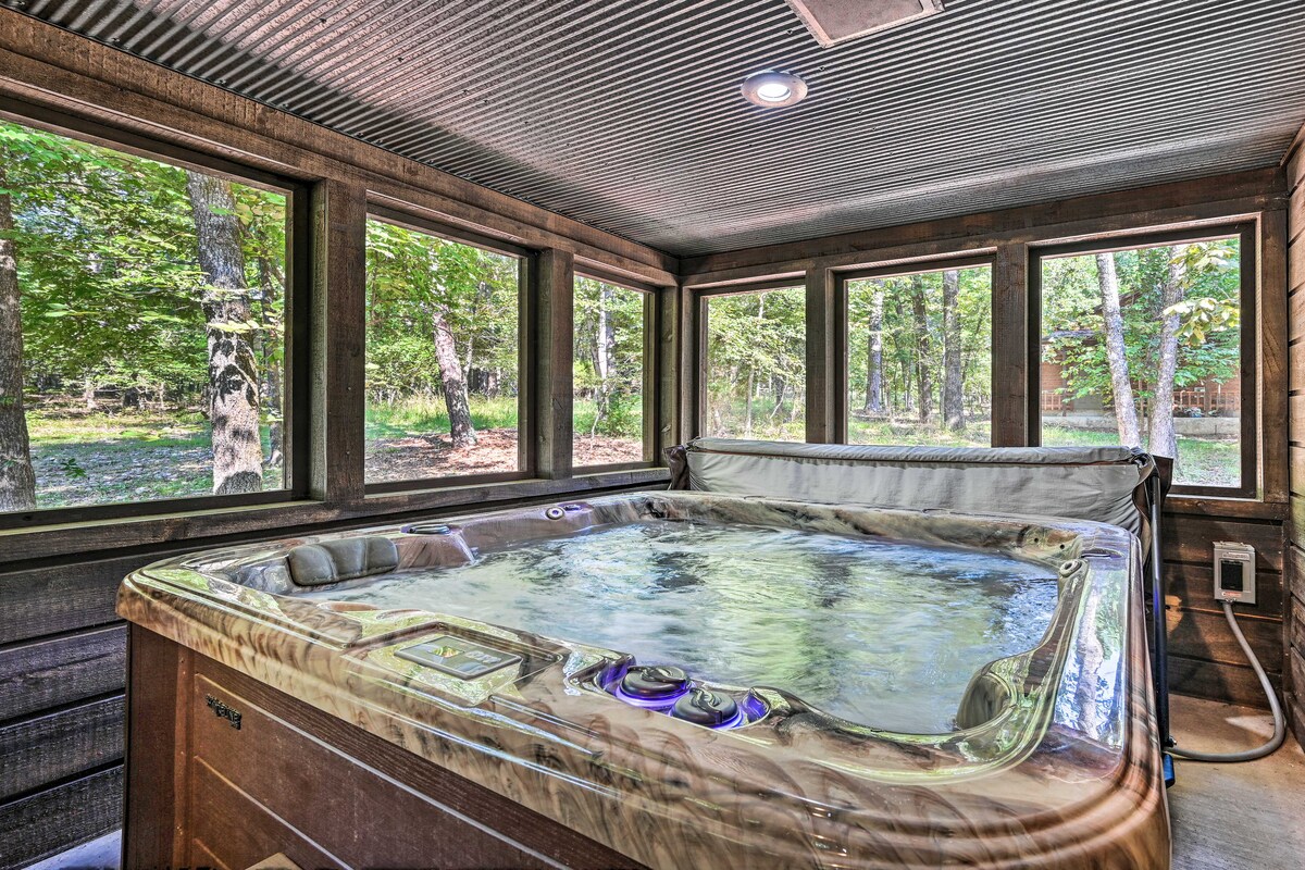 A screened-in hot tub area is surrounded by large windows that frame the natural greenery outside. The hot tub features a textured surface and controls on the side, offering a serene space for relaxation.
