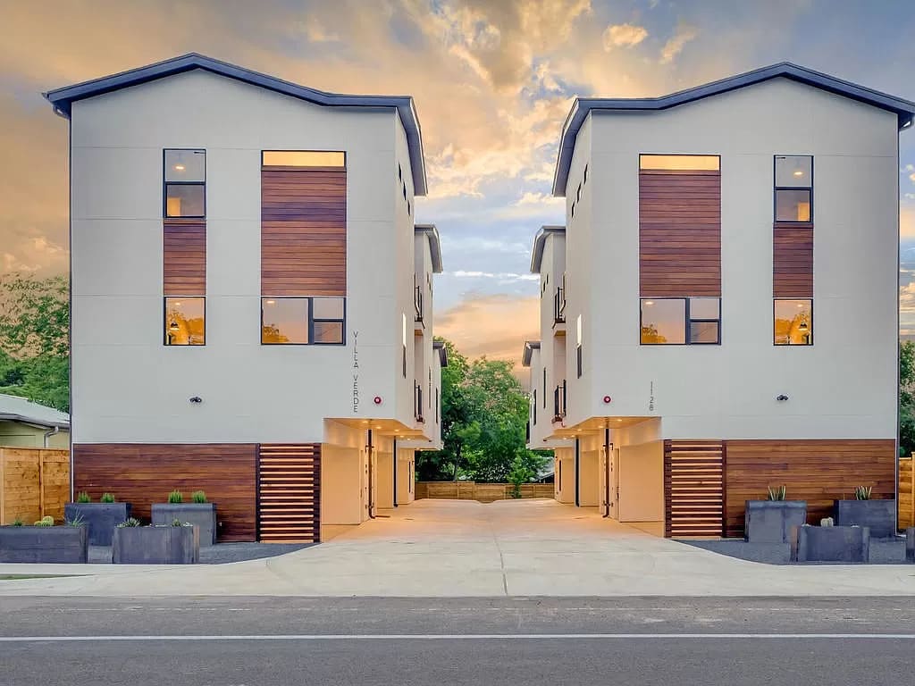 The exterior of the condo is shown, featuring two modern buildings with a combination of light-colored siding and warm wooden accents. Large windows allow natural light to illuminate the space. A spacious driveway leads to the entrances of the units, framed by decorative planters.