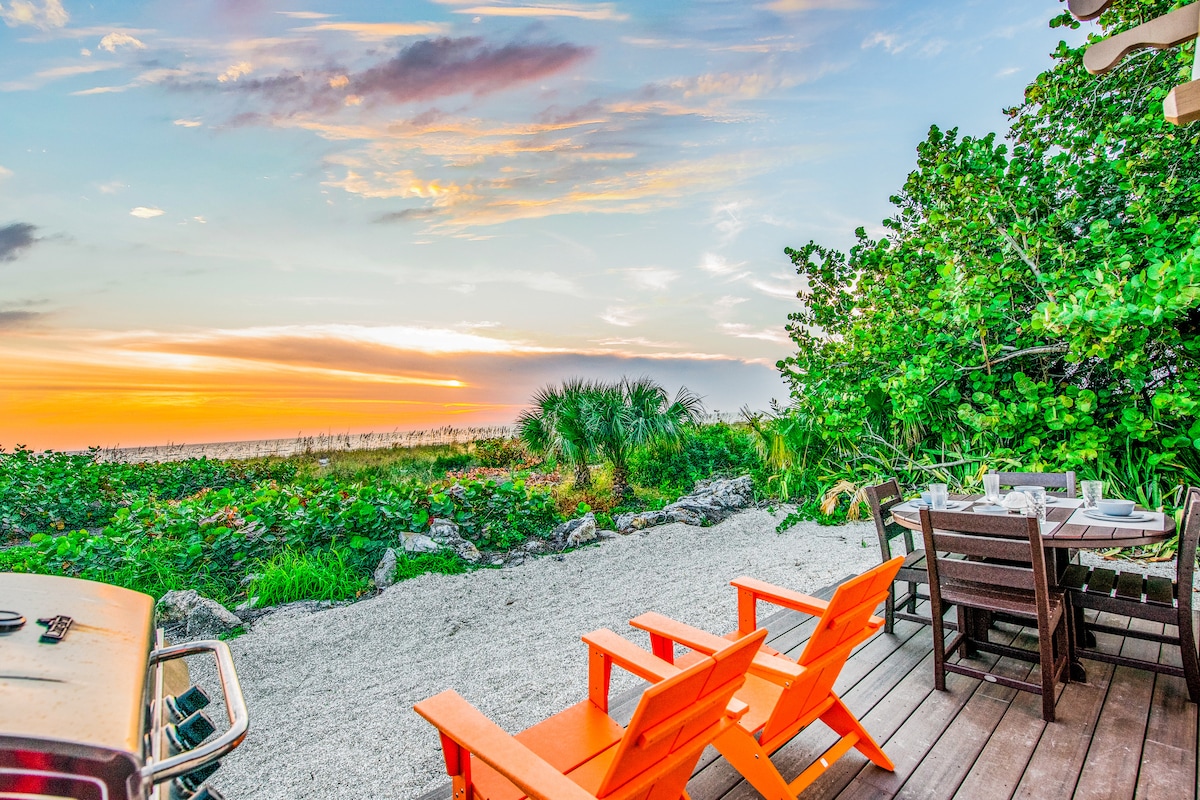 An outdoor patio features orange Adirondack chairs and a wooden dining table amidst lush greenery and sandy terrain. The scene is framed by vibrant vegetation, with a stunning sunset visible over the beach in the background.