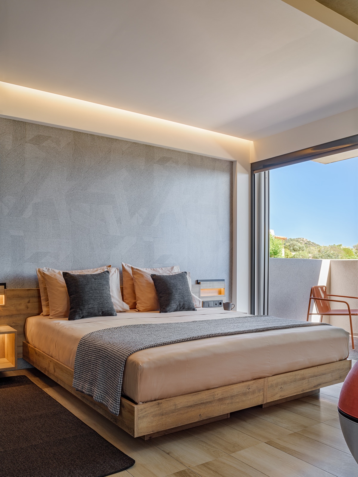 A spacious bedroom features a large bed with neutral bedding and decorative pillows. Soft lighting highlights the textured wall, while a sliding glass door opens to a private balcony, allowing natural light to fill the room. A modern reading chair and bedside tables complement the decor.