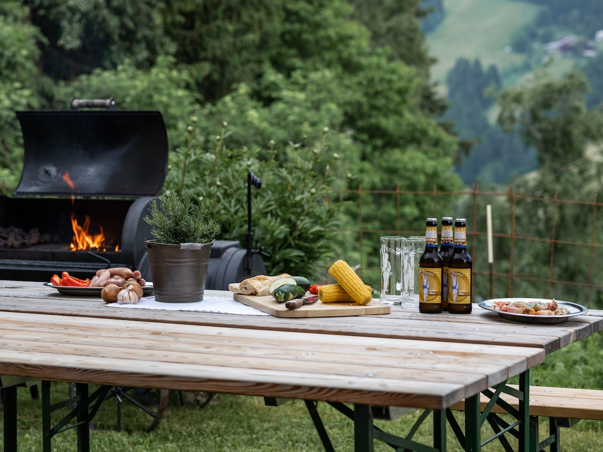 A wooden table is set outdoors, featuring a spread of grilled foods, including sausages, corn, and vegetables. Nearby, beers are placed in bottles alongside glassware. A barbecue grill is visible with smoke rising, against a backdrop of lush greenery and distant hills.