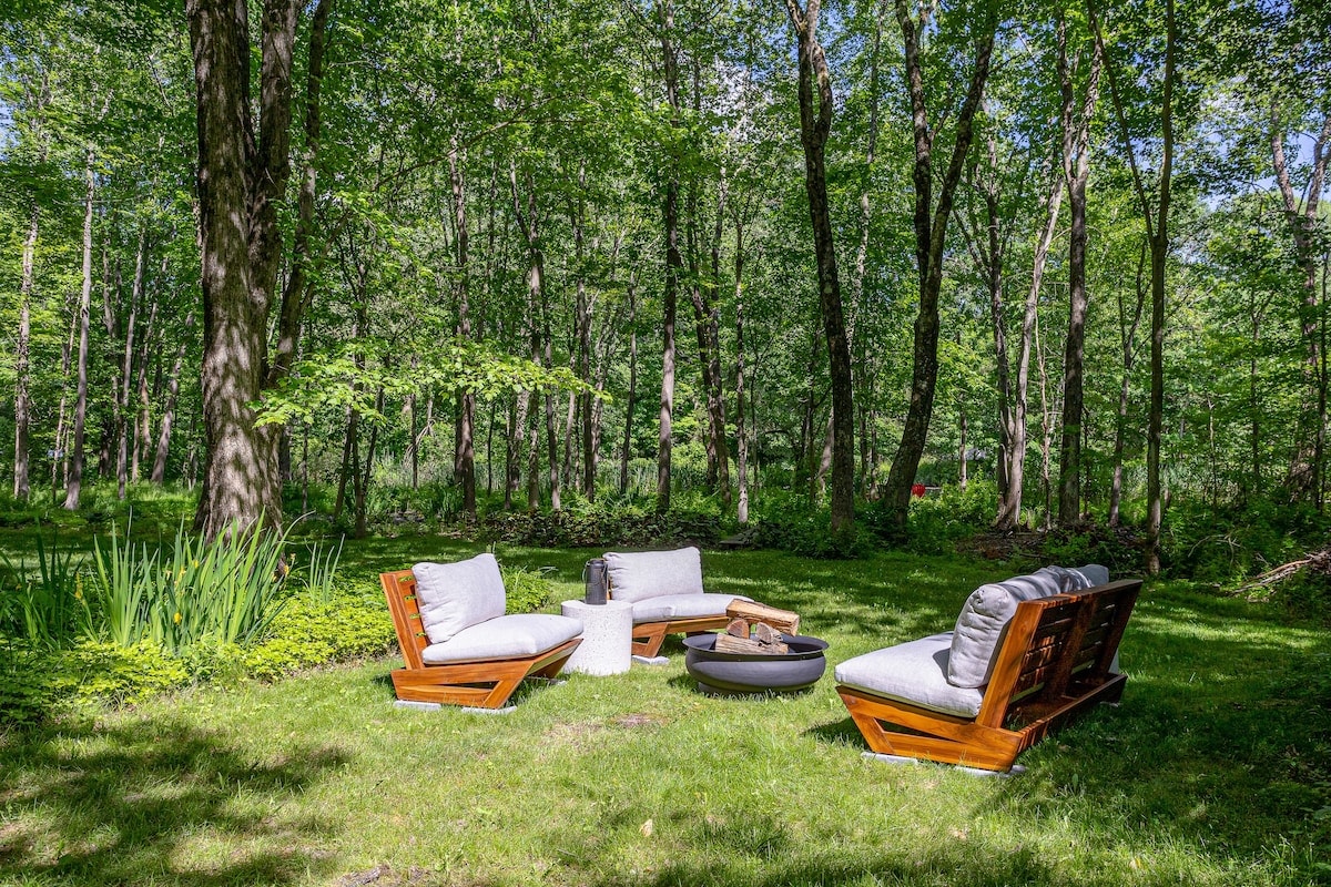 A serene outdoor seating area features four cushioned chairs and a central fire pit surrounded by lush greenery. Tall trees create a natural canopy, providing shade and privacy. The setting invites relaxation amidst the tranquil sounds of nature.