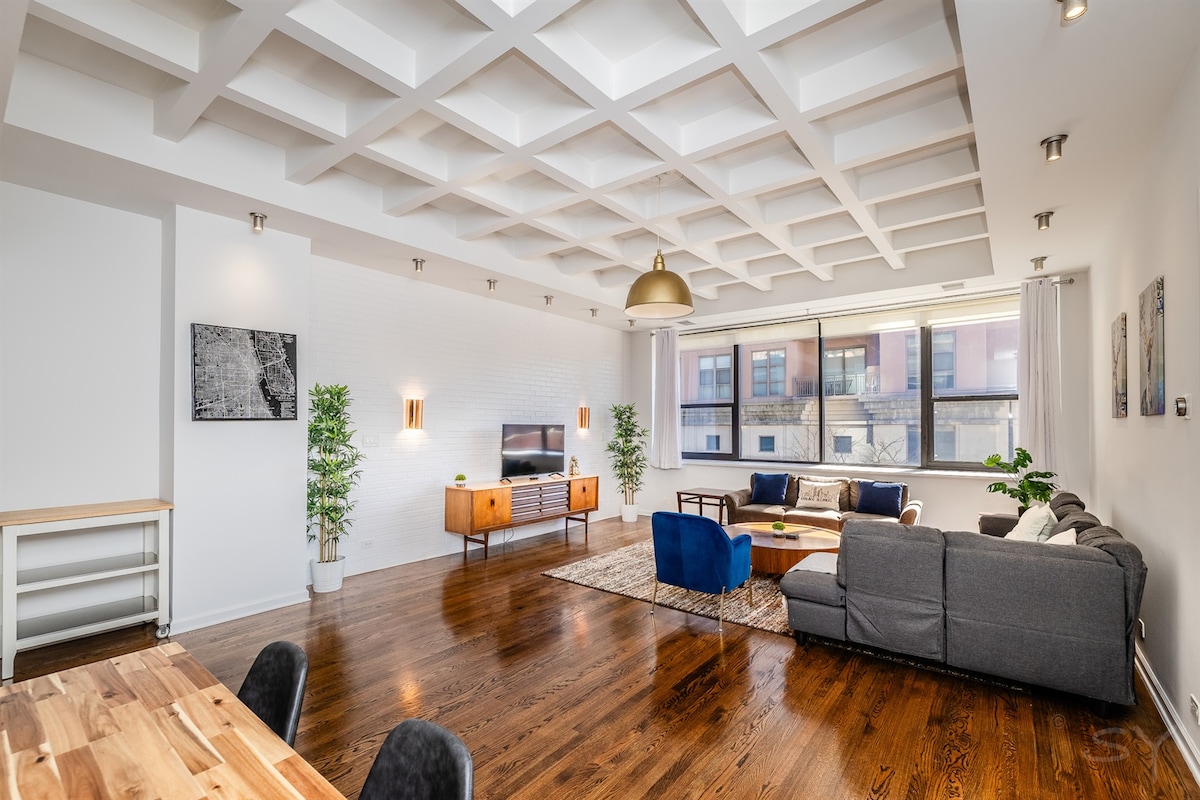 A spacious living area features a coffered ceiling and large windows allowing abundant natural light. A comfortable seating arrangement includes two sofas arranged around a central coffee table. Modern decor elements are displayed on walls and shelves, complemented by indoor plants adding a touch of greenery.