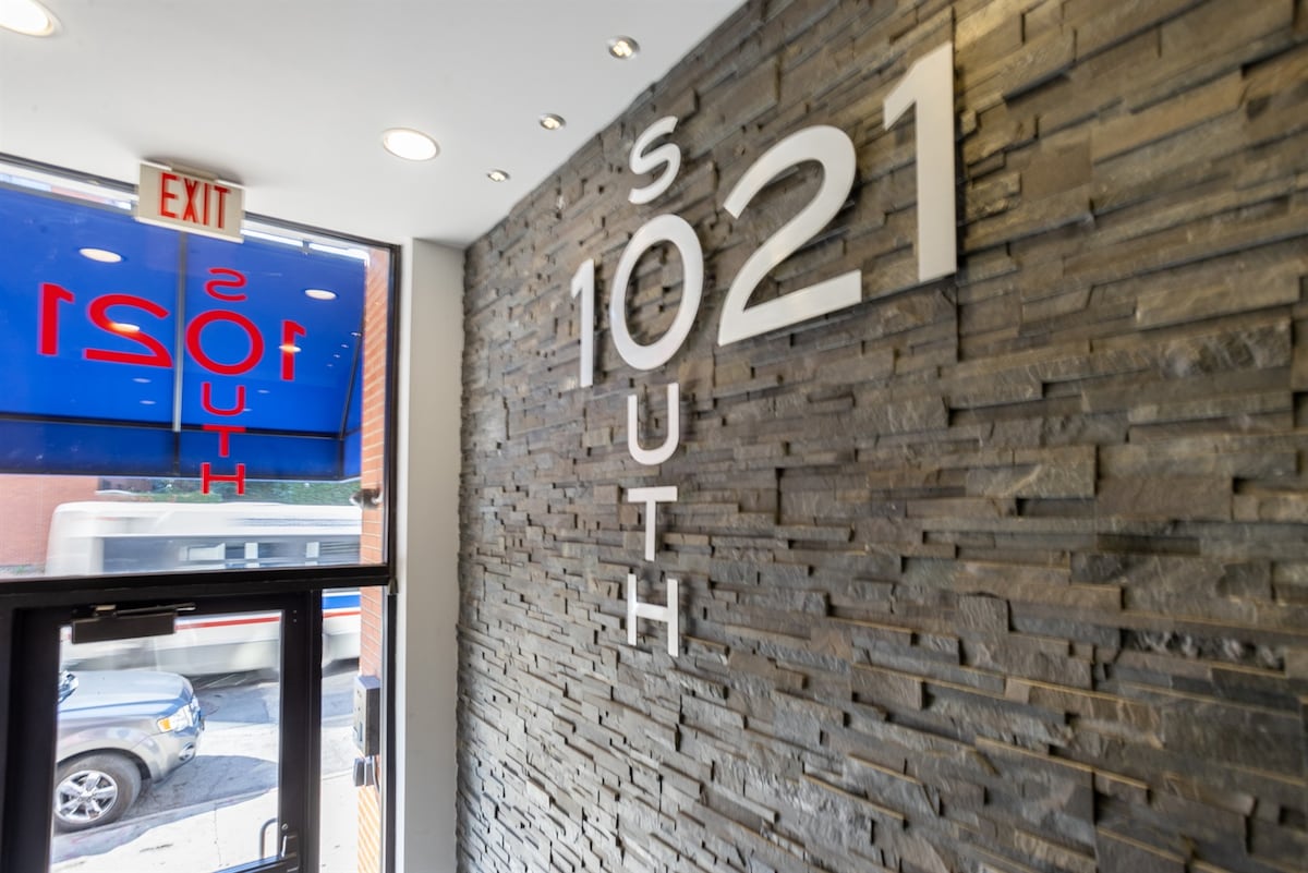 A modern entryway features a textured stone wall with large, bold numbers displaying '1021 South.' Illuminated ceiling lights highlight the contemporary design. A glass door leads outside, showcasing a glimpse of the street beyond.
